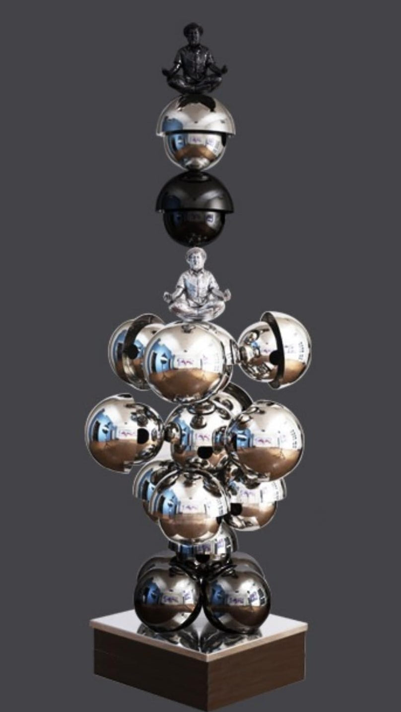 Shovin Bhattacharjee stainless steel tower sculpture featuring stacked reflective silver discs with a seated meditating figure at top.